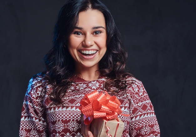 Christmas, Valentine's Day, New year. Portrait of a happy brunette girl wearing a warm sweater holding a gift box, isolated on a dark textured background.