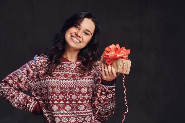 Рождество, День святого Валентина, Новый год. Портрет счастливой брюнетки в теплом свитере с подарочной коробкой, изолированной на темном текстурированном фоне.