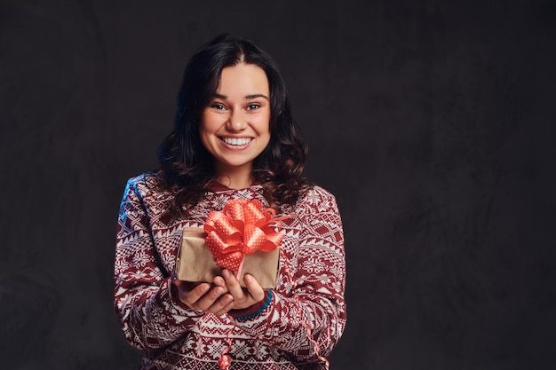 Natale, san valentino, capodanno. ritratto di una ragazza bruna felice che indossa un maglione caldo con in mano una confezione regalo, isolata su uno sfondo scuro con texture.