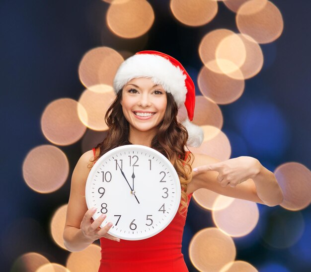 Рождество, рождество, зима, концепция счастья - улыбающаяся женщина в шляпе санта-помощника с часами, показывающими 12