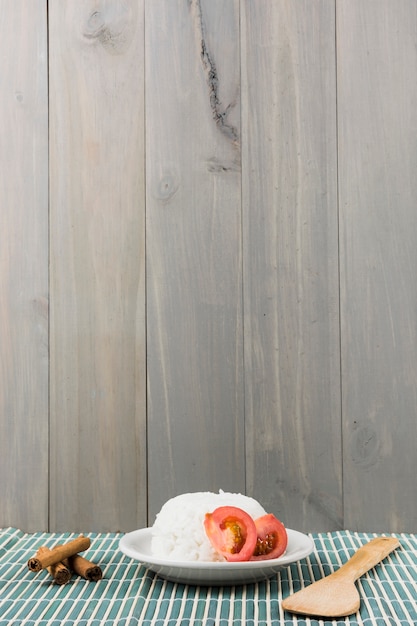 Cinnamon sticks; spatula and plate of white rice with tomato slice on placemat against wooden background