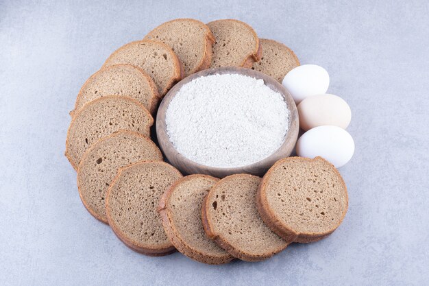 Cerchio di fette di pane nero e uova intorno a una ciotola di farina sulla superficie di marmo