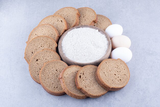 Circle of sliced black bread and eggs around a bowl of flour on marble surface