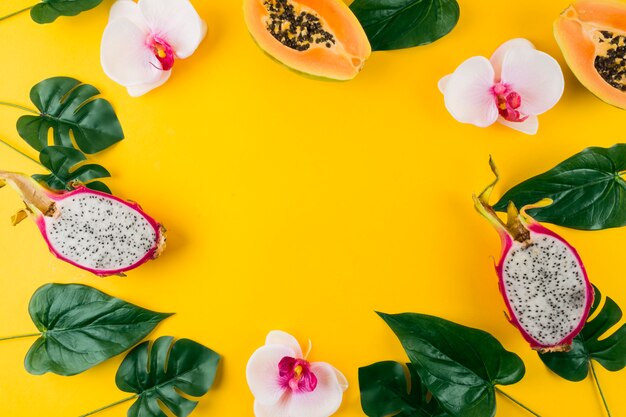 Circular frame made with artificial leaves; orchid flower; papaya and dragon fruits on yellow backdrop