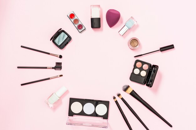 Circular frame made with makeup brushes; nail polish bottle; eyeshadow; blender on pink background