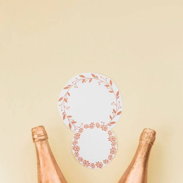 Круглая рамка с листьями и цветочным узором возле двух бутылок шампанского на бежевом фоне