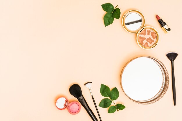 Circular mirror; compact powder; lipstick and makeup brushes with leaves on pastel background
