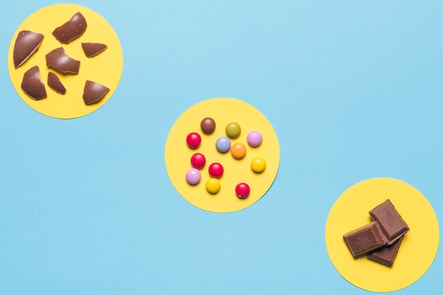 Circular yellow frame over the colorful gem candies; easter egg shells and chocolate pieces on blue background