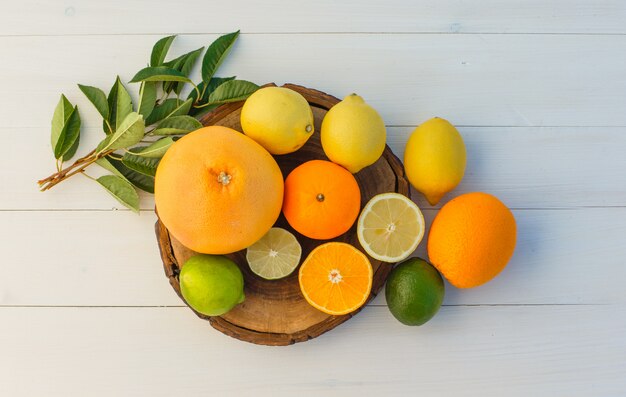 Citrus fruits with leaves on cutting board and wooden background, top view.