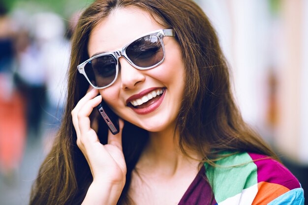 City lifestyle portrait of pretty brunette woman using smart phone, speaking and smiling, bright clothes, vintage film colors.