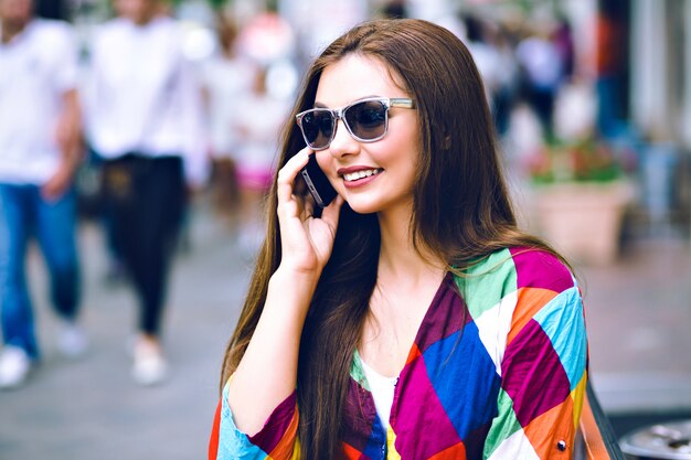 City lifestyle portrait of pretty brunette woman using smart phone, speaking and smiling, bright clothes, vintage film colors.