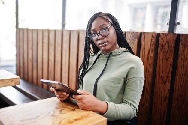 City portrait of positive young dark skinned female wearing green hoody and eyeglasses sitting with tablet