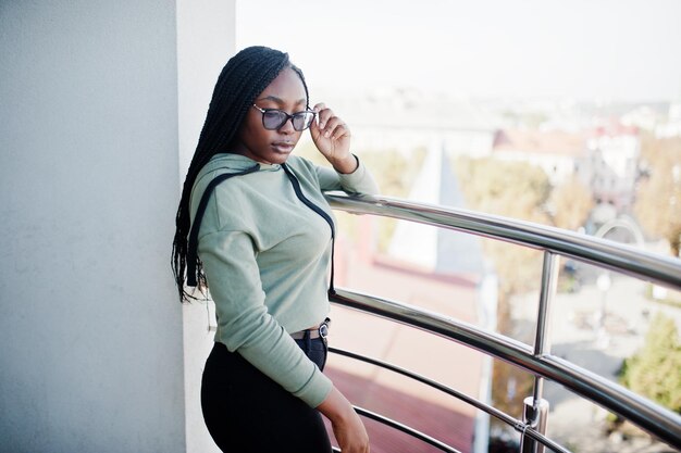 Free photo city portrait of positive young dark skinned female wearing green hoody and eyeglasses standing at balkony