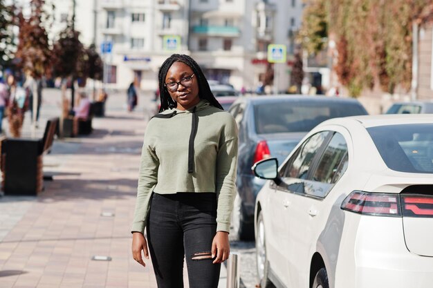 City portrait of positive young dark skinned female wearing green hoody and eyeglasses walking at car parking