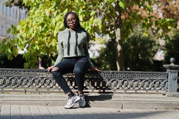 City portrait of positive young dark skinned female wearing green hoody and eyeglasses