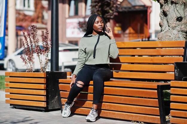 City portrait of positive young dark skinned female wearing green hoody sitting on bench