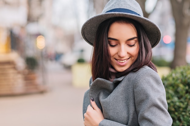 City walk time of charming young stylish woman in grey coat, hat walking on street in city. Smiling with closed eyes, expressing true positive face emotions, luxury lifestyle, elegant outlook.