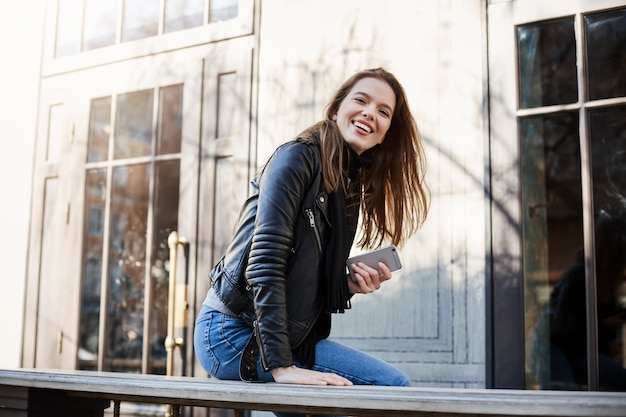 City with positive citizens. Beautiful trendy woman in leather jacket sitting near cafe, leaning on bench, looking aside