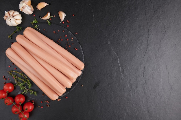 Free photo classic boiled meat pork sausages on chopping board with pepper and basil and cherry tomatoes. snack for kid. raw barbecue sausages bratwurst on a marble board. black background. top view. copy space