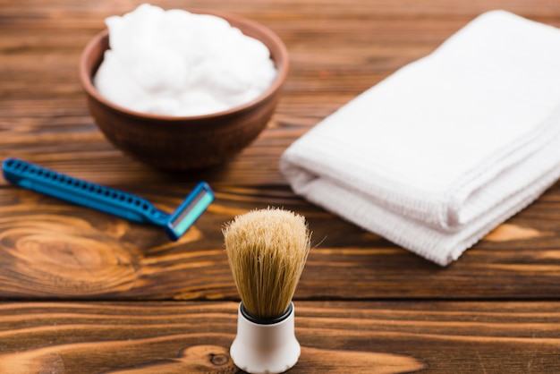 Free photo classic shaving brush in front of foam bowl; folded white napkin and razor on wooden desk