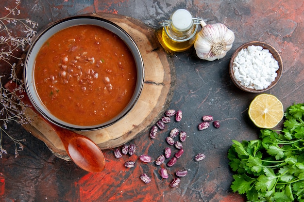 Классический томатный суп в синей миске, ложка на деревянном подносе, бутылка с маслом, чесночная соль и лимон, пучок зелени на столе смешанных цветов