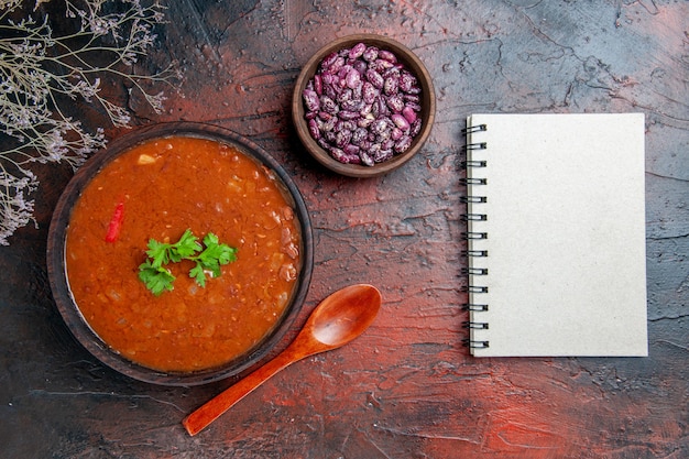 Foto gratuita zuppa di pomodoro classica in una ciotola marrone fagioli e cucchiaio accanto al taccuino sulla tavola di colori misti