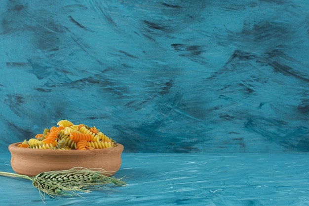 Clay bowl of colorful raw fusilli pasta on blue background. 