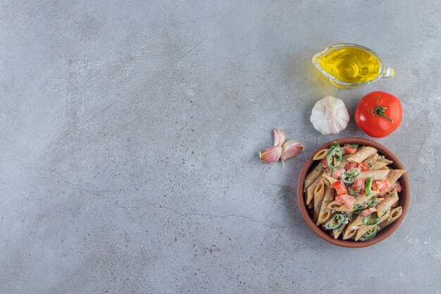 Clay bowl of delicious penne with chopped vegetables and oil on stone background.