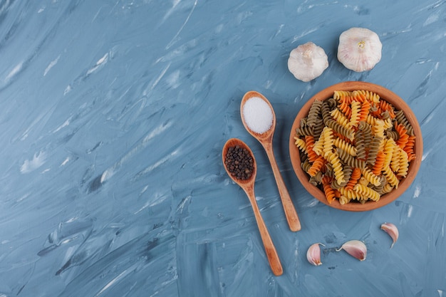 Free photo a clay bowl of multi colored raw pasta with garlics on a blue table.