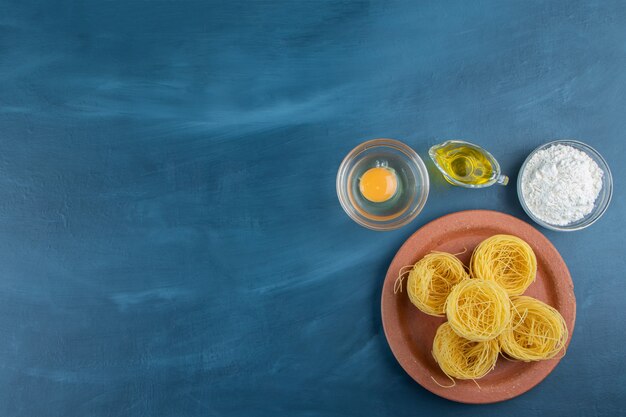 A clay plate of raw dry nest pasta with raw egg and oil on a dark-blue background . 