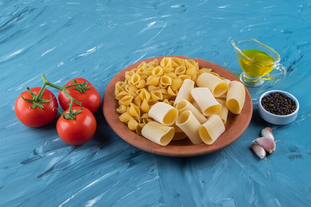 A clay plate of uncooked pasta with oil and fresh red tomatoes on a blue surface .