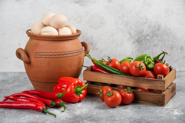 Clay pot full of raw chicken eggs and fresh vegetables on marble.