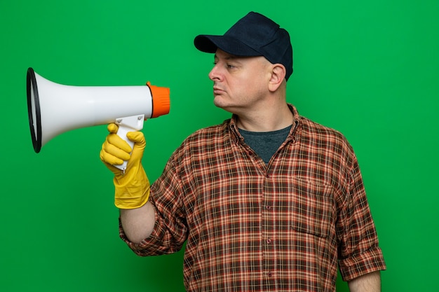 Бесплатное фото Уборщик в клетчатой рубашке и кепке в резиновых перчатках с мегафоном смотрит в сторону с уверенным выражением лица, стоя на зеленом фоне