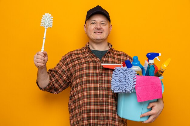 Уборщик в клетчатой рубашке и кепке держит ведро с инструментами для чистки и щеткой для чистки, глядя в камеру, уверенно улыбаясь, готовый к уборке, стоя на оранжевом фоне