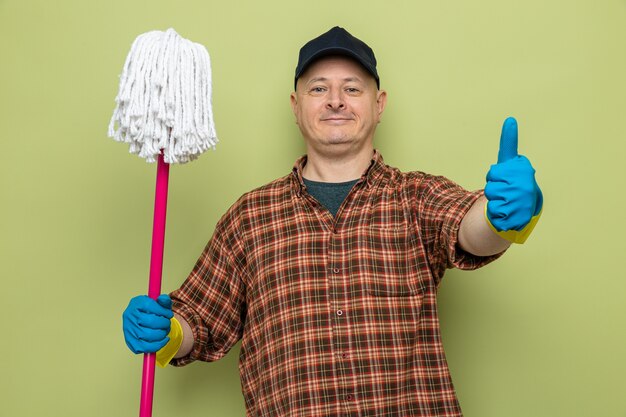 Cleaning man in plaid shirt and cap wearing rubber gloves holding mop looking at camera with confident smile on face showing thumb up standing over green background