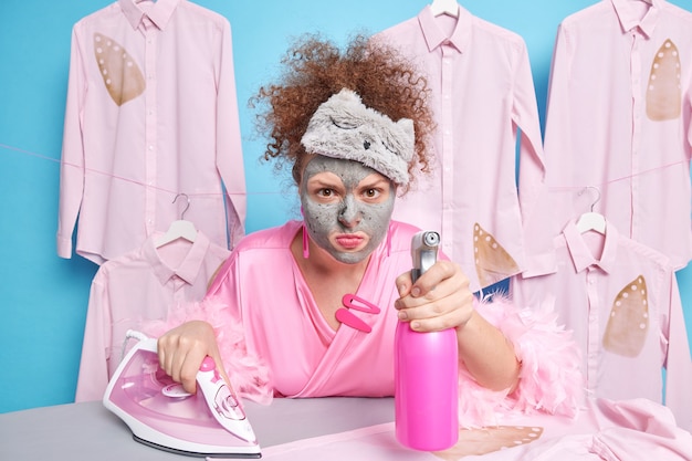 Cleaning service hygiene and housework concept. Scrupulous curly haired woman maid looks annoyed  applies facial clay mask holds cleaning detergent sprays in room busy ironing clothes