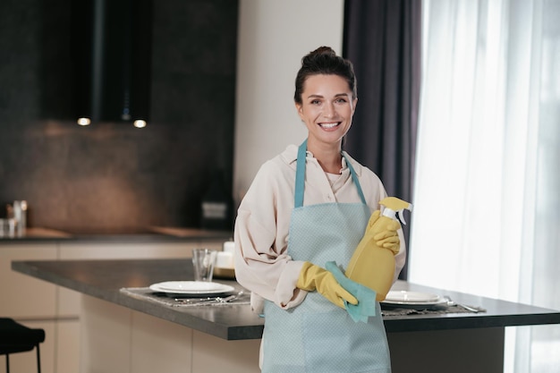 Бесплатное фото Уборка кухни. улыбающаяся молодая домохозяйка держит в руках дезинфицирующий спрей