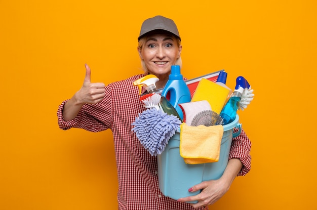 Cleaning woman in plaid shirt and cap holding bucket with cleaning tools looking at camera happy and positive smiling cheerfully showing thumbs up standing over orange background