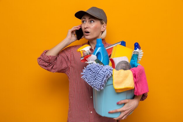 Donna delle pulizie in camicia a quadri e berretto che tiene il secchio con strumenti per la pulizia che sembra sorpresa mentre parla al telefono cellulare in piedi su sfondo arancione