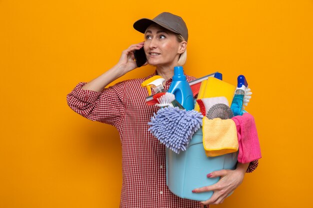 Cleaning woman in plaid shirt and cap holding bucket with cleaning tools smiling confident while talking on mobile phone