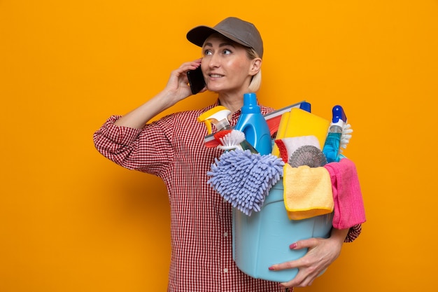 Cleaning woman in plaid shirt and cap holding bucket with cleaning tools smiling confident while talking on mobile phone