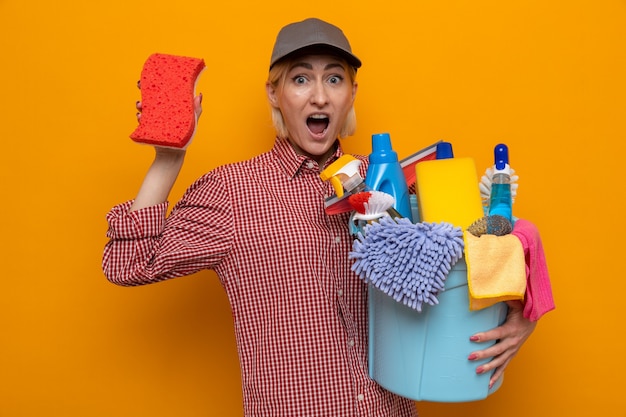 Уборщица в клетчатой рубашке и кепке держит губку и ведро с чистящими средствами, глядя в камеру изумленно и удивленно, стоя на оранжевом фоне
