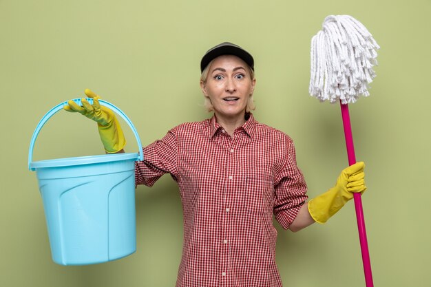 Cleaning woman in plaid shirt and cap wearing rubber gloves holding bucket and mop looking smiling happy and positive