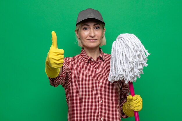 Уборщица в клетчатой рубашке и кепке в резиновых перчатках держит швабру, глядя в камеру, уверенно улыбаясь, показывая пальцы вверх, стоя на зеленом фоне
