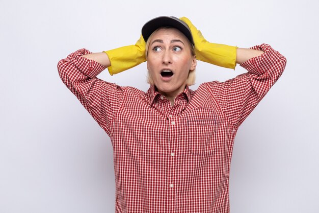 Cleaning woman in plaid shirt and cap wearing rubber gloves looking aside amazed and surprised holding hands on her head