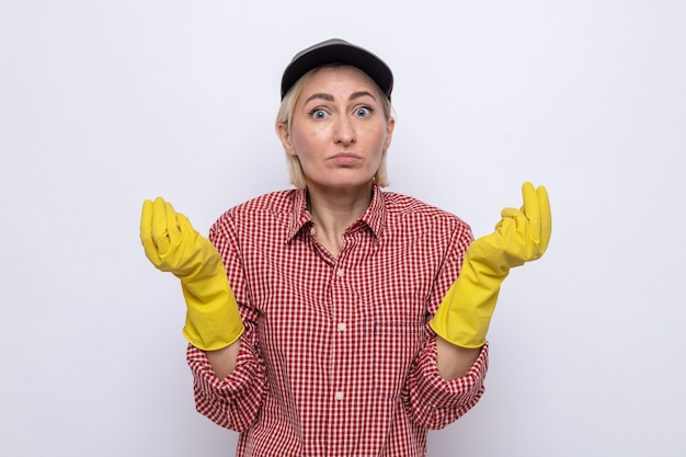 Cleaning woman in plaid shirt and cap wearing rubber gloves looking at camera confused rubbing fingers making money gesture standing over white background