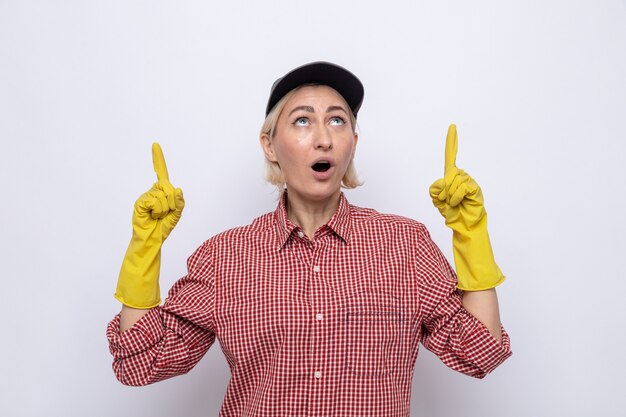 Cleaning woman in plaid shirt and cap wearing rubber gloves looking up surprised pointing with index fingers up