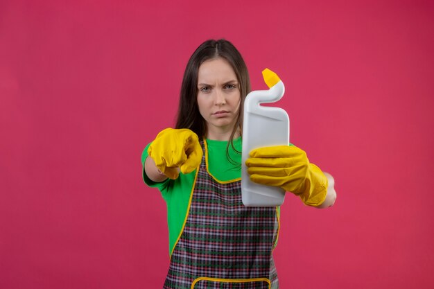 чистящая молодая женщина в униформе в перчатках держит чистящее средство, показывая вам жест на изолированной розовой стене
