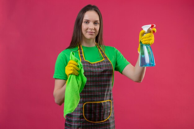  cleaning young woman wearing uniform in gloves holding cleaning spray and rag on isolated pink wall