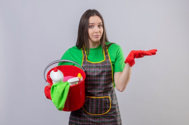 Pulizia giovane donna che indossa l'uniforme in guanti rossi che tengono gli strumenti di pulizia alzando la mano sul muro bianco isolato